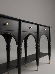 A black wooden console table with three drawers, arched legs, a lower shelf, and brass-finished drawer knobs and leg caps, photographed against a neutral gray studio background.