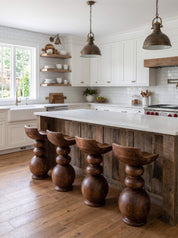 A rustic farmhouse kitchen island with a white stone countertop and four carved dark wood bar stools, set in a bright kitchen with white cabinets, wood floors, and hanging pendant lights.