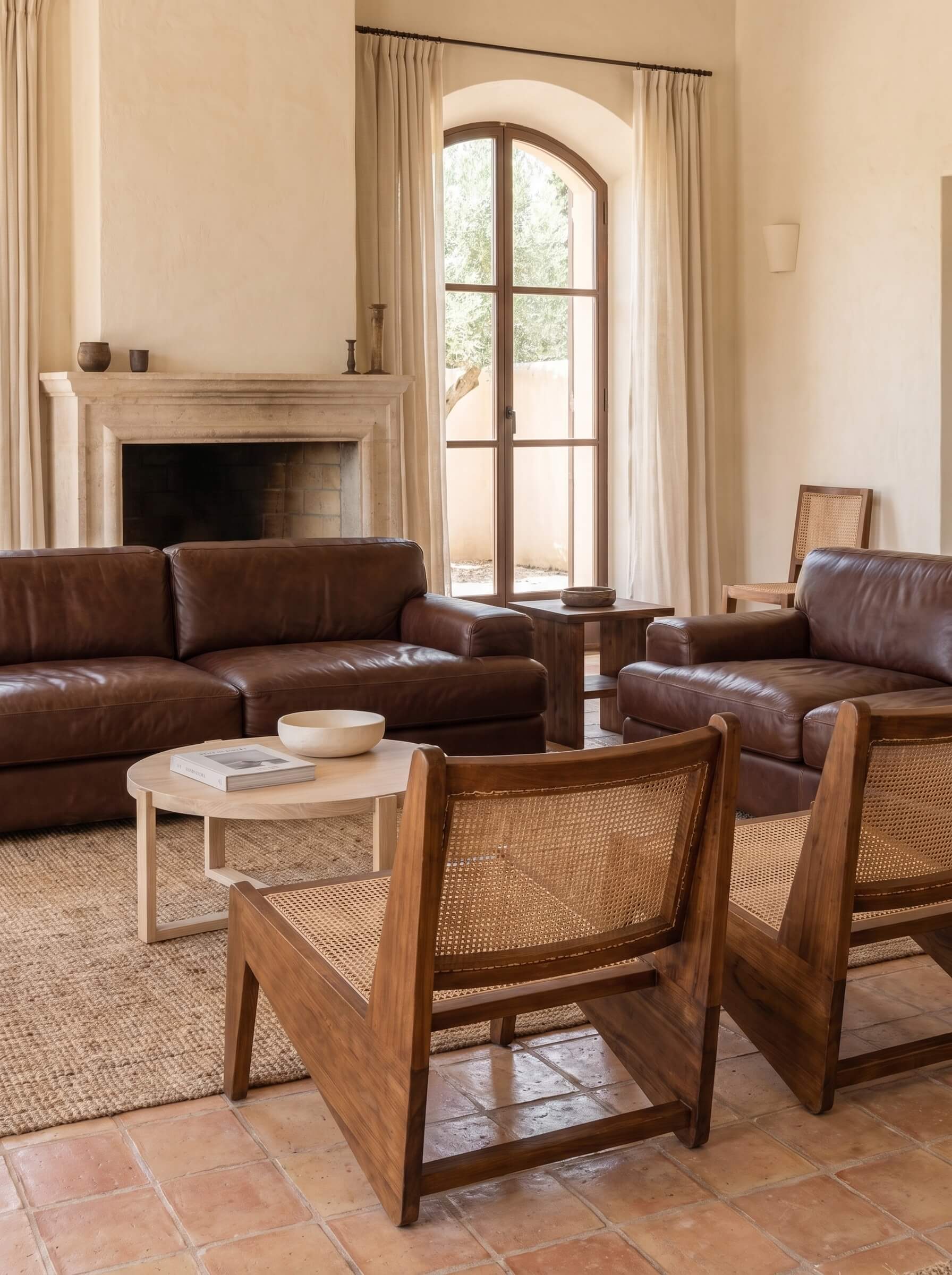 A styled living room featuring dark brown leather sofas, two wooden armchairs with woven cane backs, a round light wood coffee table, and a large arched window with cream drapes.