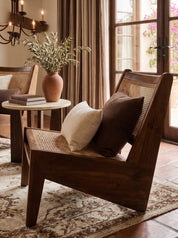 A rustic wooden armchair with woven cane back and seat, styled with a cream pillow and a dark brown accent pillow in a warm, sunlit room.