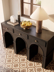 A dark wood console table with three arched legs and small drawer pulls, styled in a room with patterned tile flooring, a window, a table lamp, and decorative objects on top.