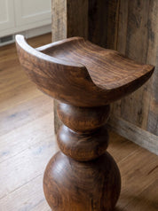 A rustic wooden pedestal stool with a carved seat, shown in a warm indoor setting with wood flooring and a wooden wall panel background.