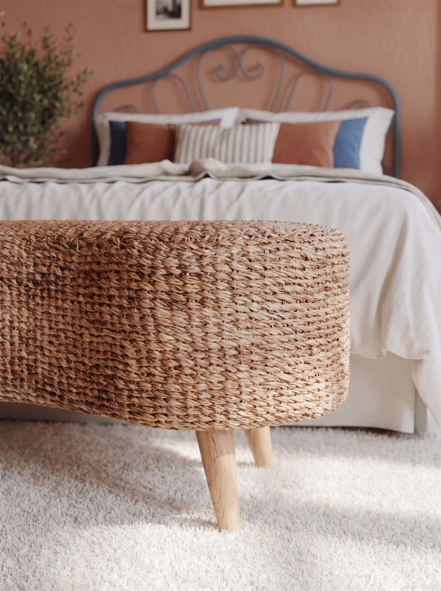 A woven natural-fiber ottoman bench with light wood legs in a softly lit bedroom, positioned in front of a bed with neutral-toned bedding and pillows.
