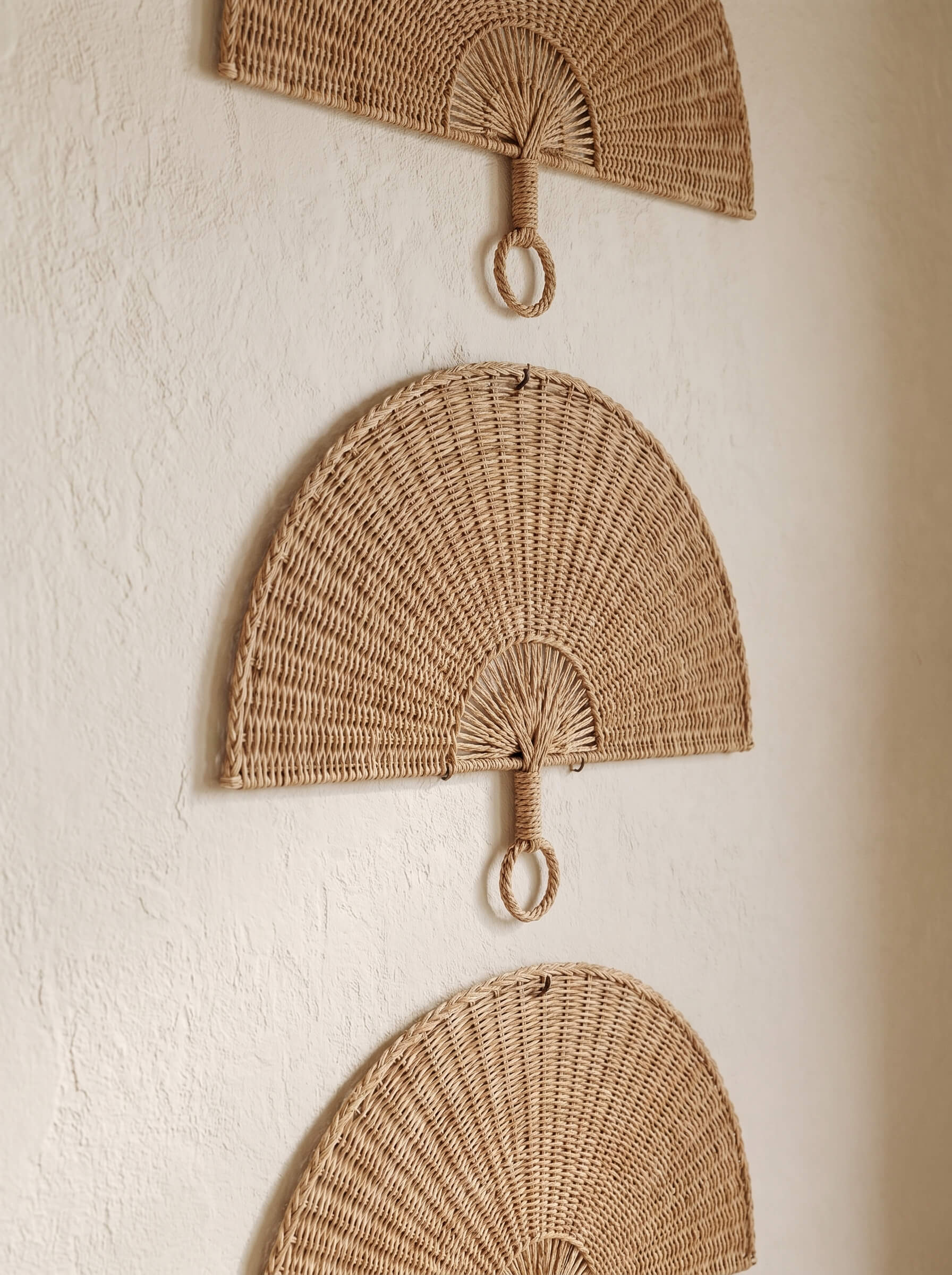 Three woven wall-mounted fans in a natural tan color, arranged vertically on a textured off-white wall.