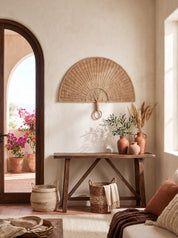 A styled bohemian interior scene featuring a natural woven wicker wall fan mounted on a textured beige plaster wall above a rustic wooden console table. The table is decorated with terracotta vases, dried pampas grass, and greenery, with matching woven baskets on the floor.