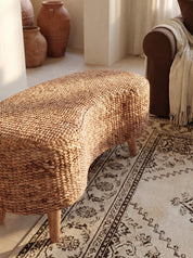 A curved woven rattan bench with light wood legs, styled in a warm, sunlit room on a patterned rug next to a brown armchair with a cream throw blanket.