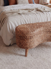 A woven natural-fiber upholstered bench with tapered wooden legs positioned at the foot of a bed in a softly styled bedroom.