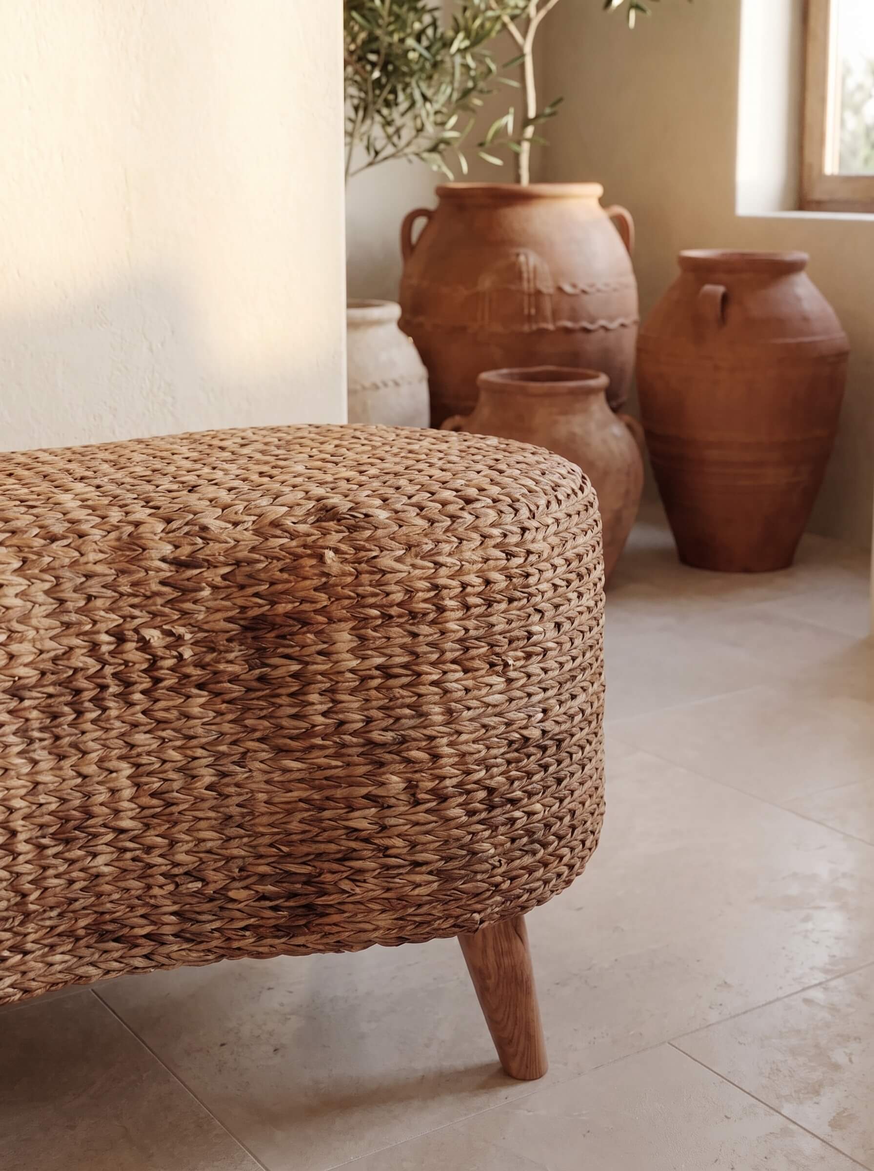A woven natural-fiber ottoman or bench with a rounded rectangular shape and short wooden legs, shown in a softly lit interior with large terracotta vases and a plant in the background.
