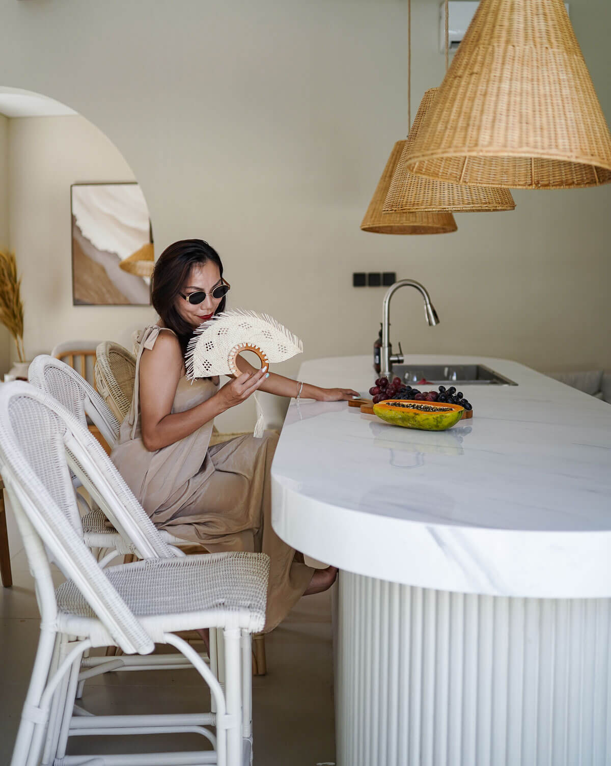 Woman sitting at a kitchen island with a plate of grapes and a fan, wearing sunglasses.