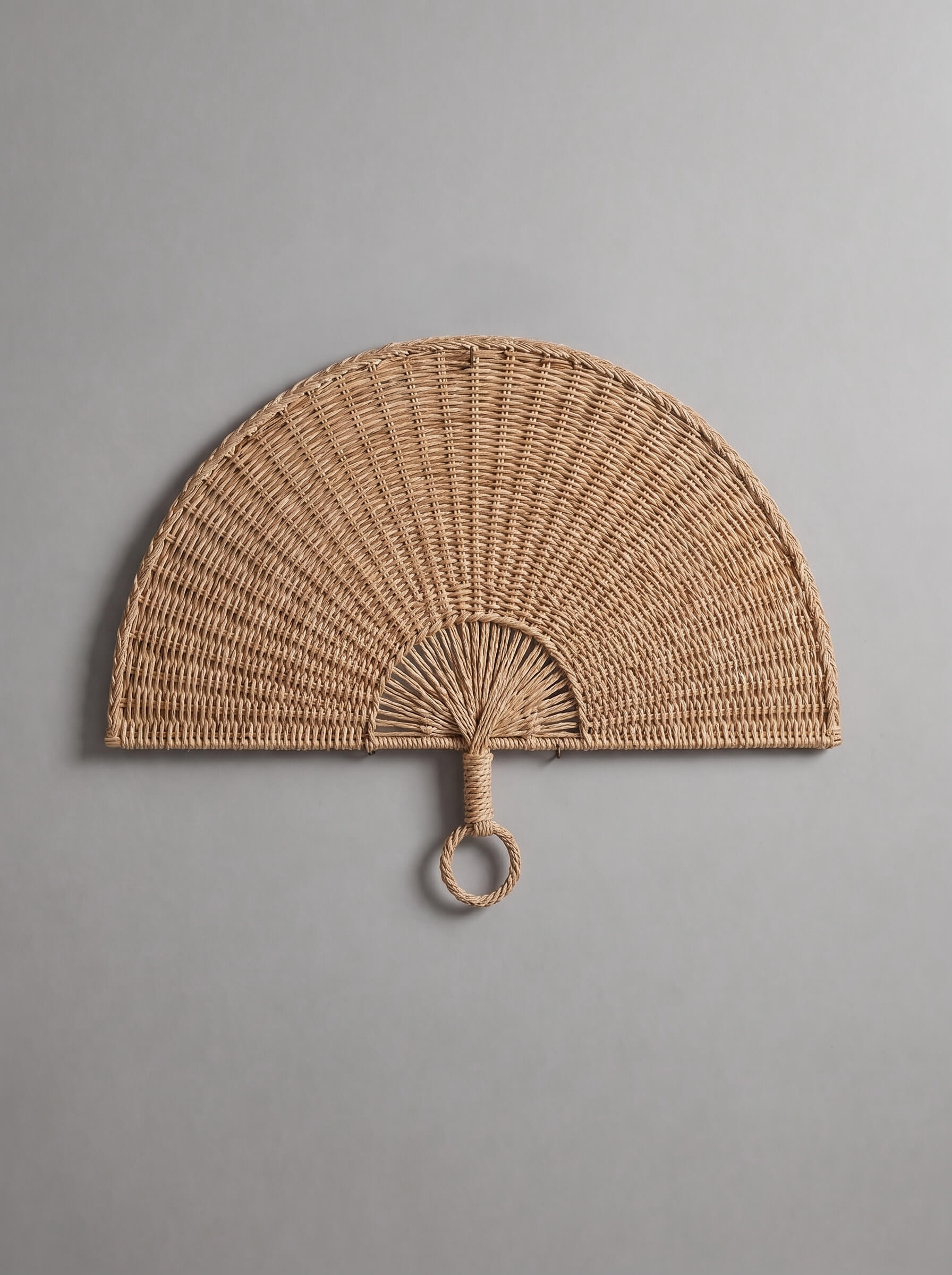 A semi-circular woven rattan wall fan with a hanging loop, shown against a solid gray background.