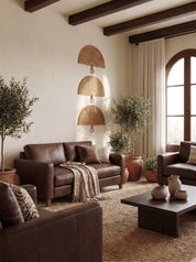 A warm, rustic living room featuring a dark brown leather sofa and matching armchair, a dark wood coffee table, a textured beige rug, and three woven wicker wall fans mounted on a cream-colored wall.